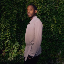 A person with short hair stands outdoors against a lush green wall of foliage. 的y are wearing a light-colored, long-sleeve shirt and are looking slightly to the side with a calm 表达式. 的 lighting casts a soft shadow, creating a warm, natural ambiance.